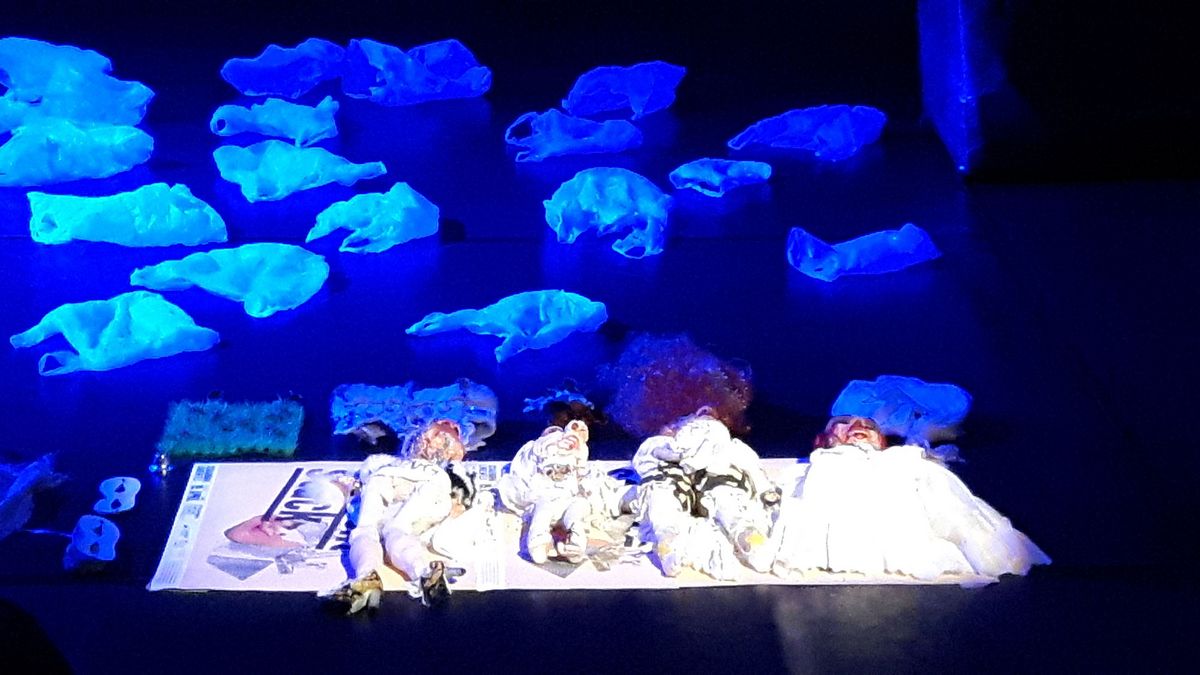 Puppen auf dem Bühnenboden, dahinter Tüten in blauem Licht