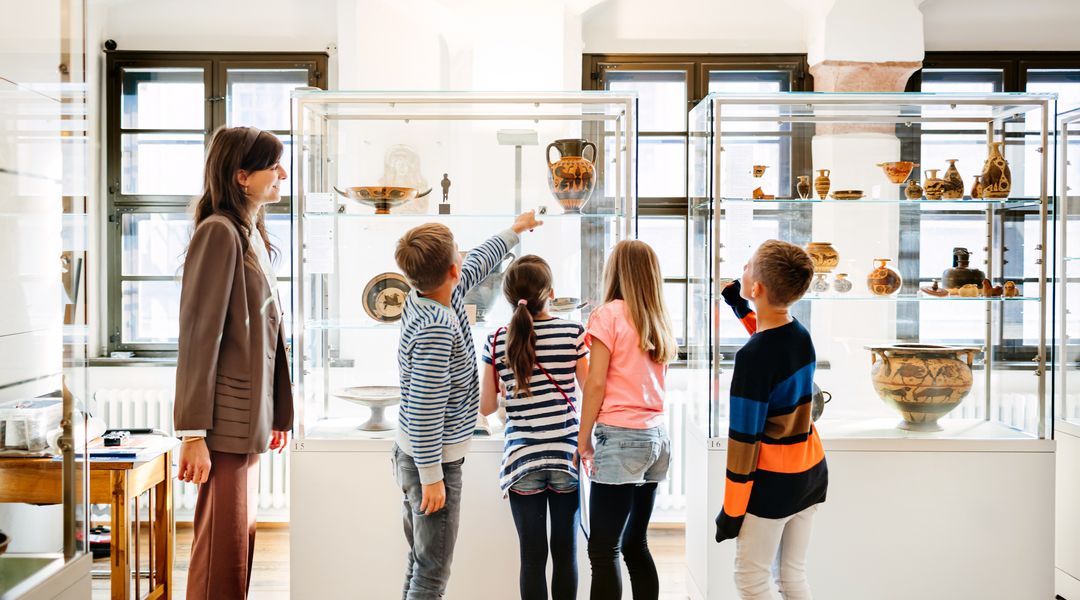 Kinder stehen zusammen mit einer Mitarbeiterin vor einer Vitrine im Antikenmuseum und schauen sich die Ausstellungsstücke an.