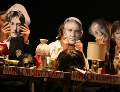 Bild einer Performance; drei Performer sitzen an einem Tisch und halten sich schwarz-weiße Pappköpfe prominenter Personen vors Gesicht, darunter Angela Merkel, Mao Zedong und John Lennon