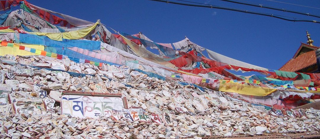 Mauer aus Steinen und Steinplatten mit Gebetssprüchen in tibetischer Schrift, darüber bunte tibetische Gebetsfahnen, Ost-Tibet, 2006 Fotograf: Franz Xaver Erhard 