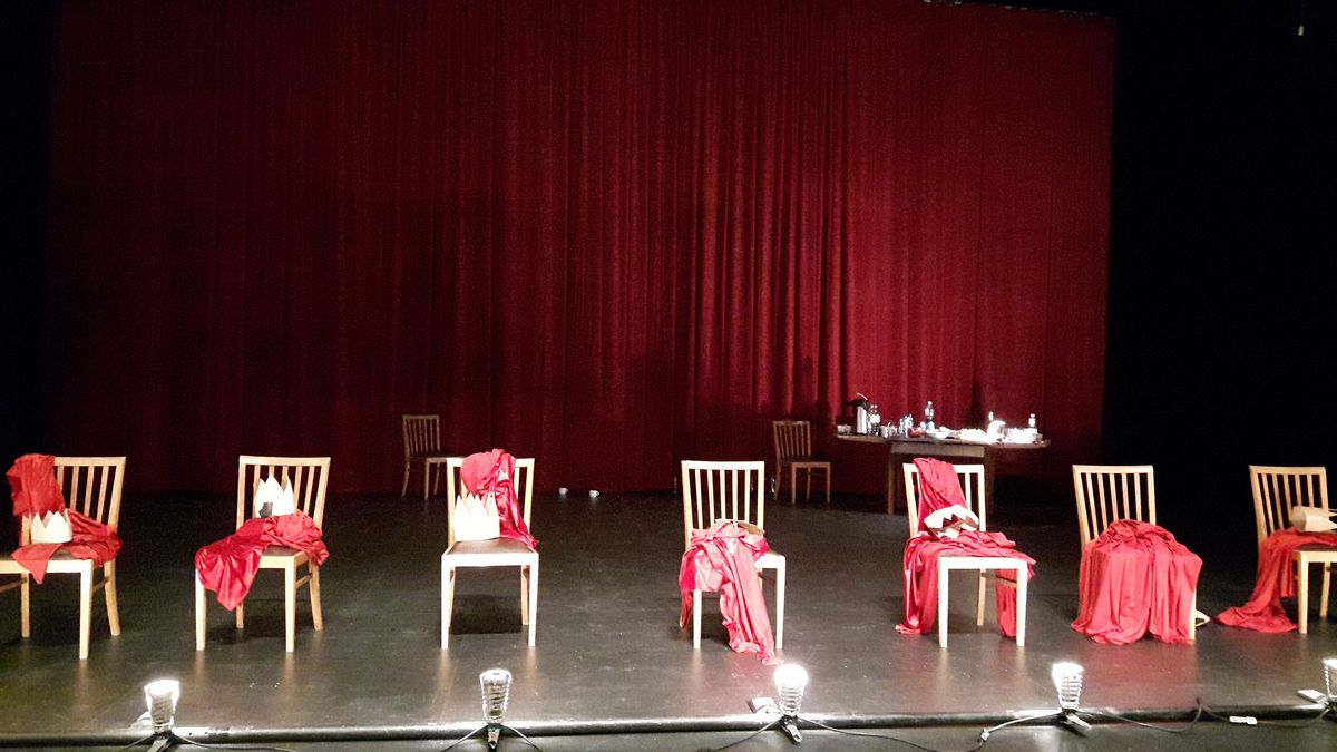 Auf dem Proszenium einer Theaterbühne stehen mehrere Holzstühle, auf denen rote Umhänge und Papierkronen liegen; im Hintergrund ein roter Theatervorhang