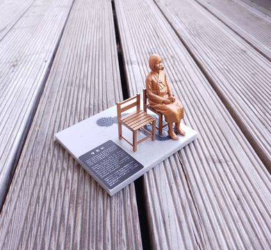 Zu sehen ist eine Miniatur des Denkmals "Trostfrauen" in goldener Farbe: Eine ins Leere blickende Frau sitzt auf einem Stuhl, der Stuhl danaben ist leer.