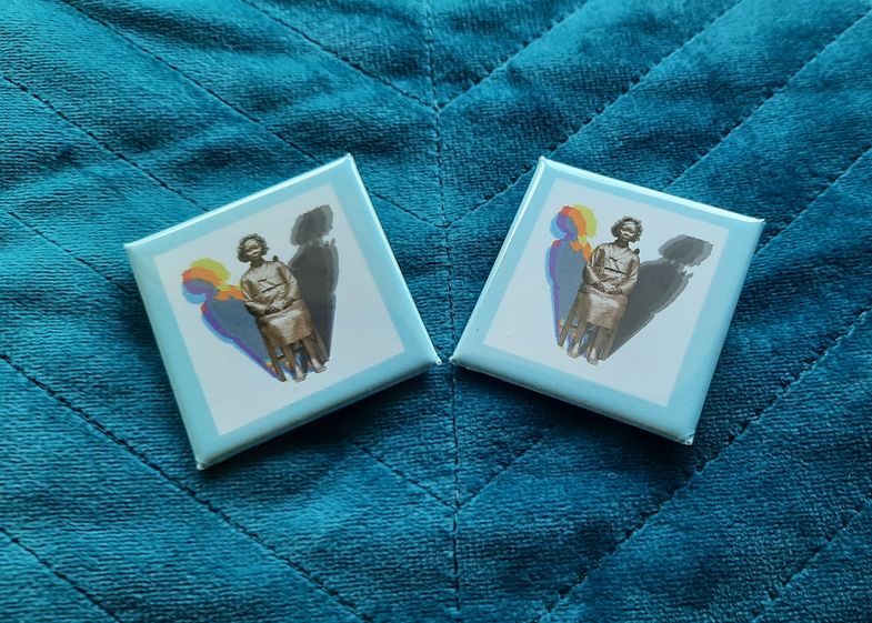 Das Bild zeigt zwei Buttons mit einem Bild der Friedensstatue auf einem blauen Stoffuntergrund.