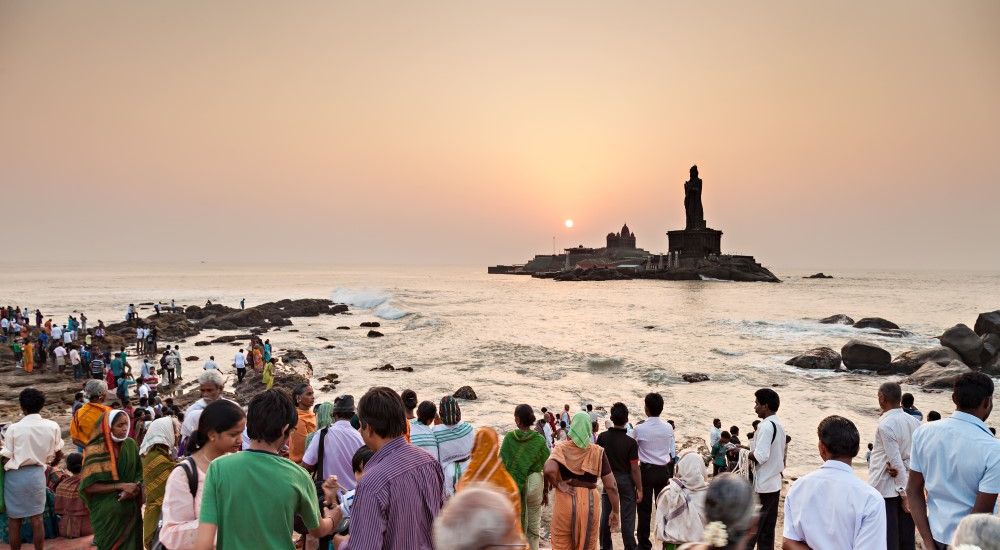 Zahlreiche Menschen stehen am Strand und richten ihren Blick auf eine kleine Insel mit einer großen Statue