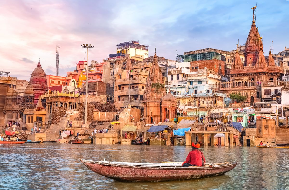 enlarge the image: In the center of the picture, you can see the city of Varanasi. In the right foreground, there is a man sitting in a boat. The photo is taken from © Adobe Stock and was shot by Roop Dey. 