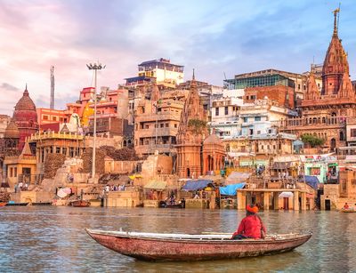 In der Mitte des Bildes ist die Stadt Varanasi zu sehen. Im rechten Bildvordergrund sitzt ein Mann in einem Boot. Das Foto stammt aus © Adobe Stock und wurde von Roop Dey aufgenommen. 