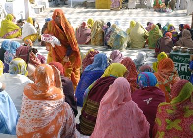 Viele Frauen sitzen in bunten Saris zusammen. Sie sind von Hinten fotografiert. Eine alte Frau geht zwischen ihnen hindurch mit einer Tüte in der Hand.