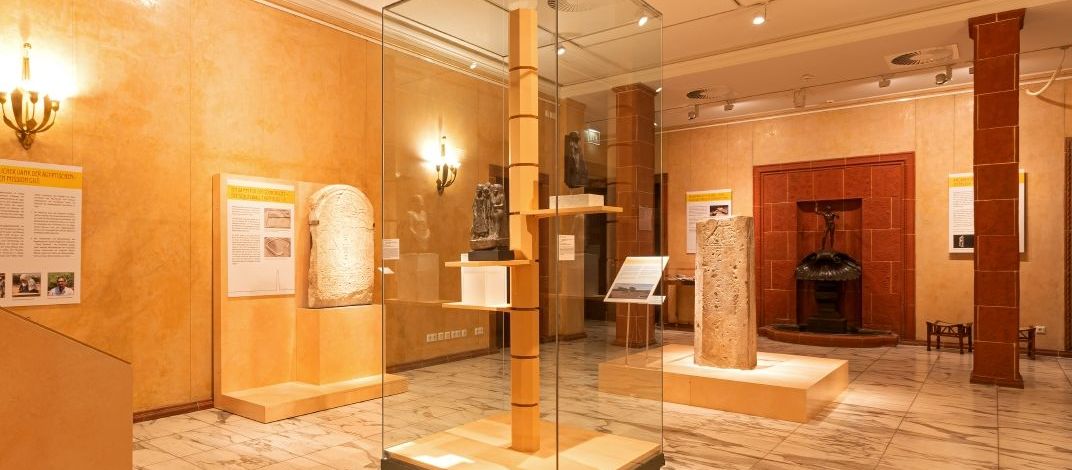 Blick in den Ausstellungsraum mit deckenhoher Vitrine in der Mitte, in welcher Skulpturen ausgestellt sind