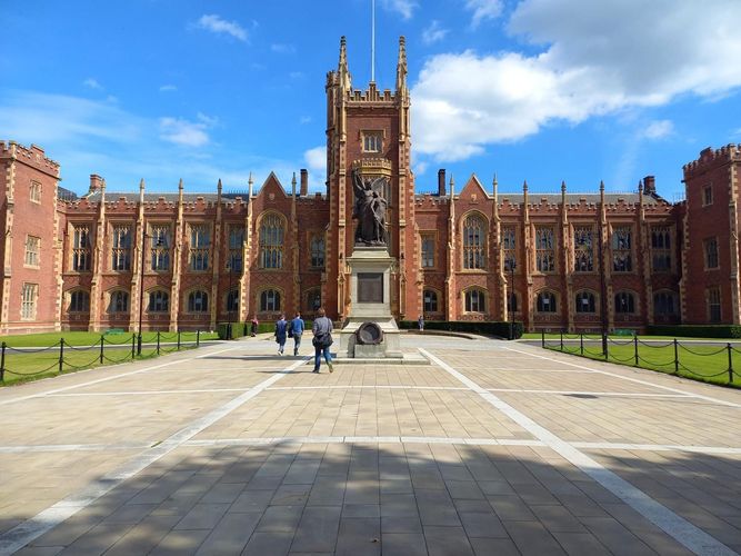 Auf dem Foto ist die Frontansicht der Queen's University Belfast, Nordirland zu sehen. Es handelt sich um einen rötlichen Backsteinbau in viktorianischem Stil, auf den ein breiter WEg hin führt. Vor dem Gebäude befindet sich eine Statue. Das Foto ist bei Sonne und strahlendblauem Himmel aufgenommen worden.