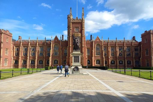 Auf dem Foto ist die Frontansicht der Queen's University Belfast, Nordirland zu sehen. Es handelt sich um einen rötlichen Backsteinbau in viktorianischem Stil, auf den ein breiter WEg hin führt. Vor dem Gebäude befindet sich eine Statue. Das Foto ist bei Sonne und strahlendblauem Himmel aufgenommen worden.