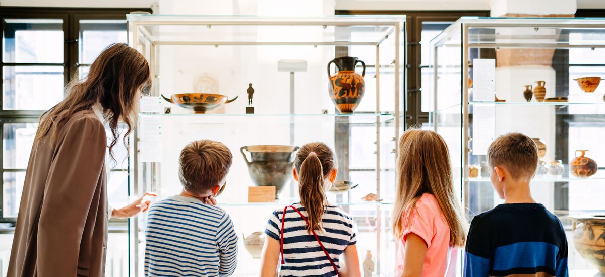 Eine Kindergruppe und eine Mitarbeiterin des Museums stehen im Museum vor einer Vitrine und betrachten die Objekte.