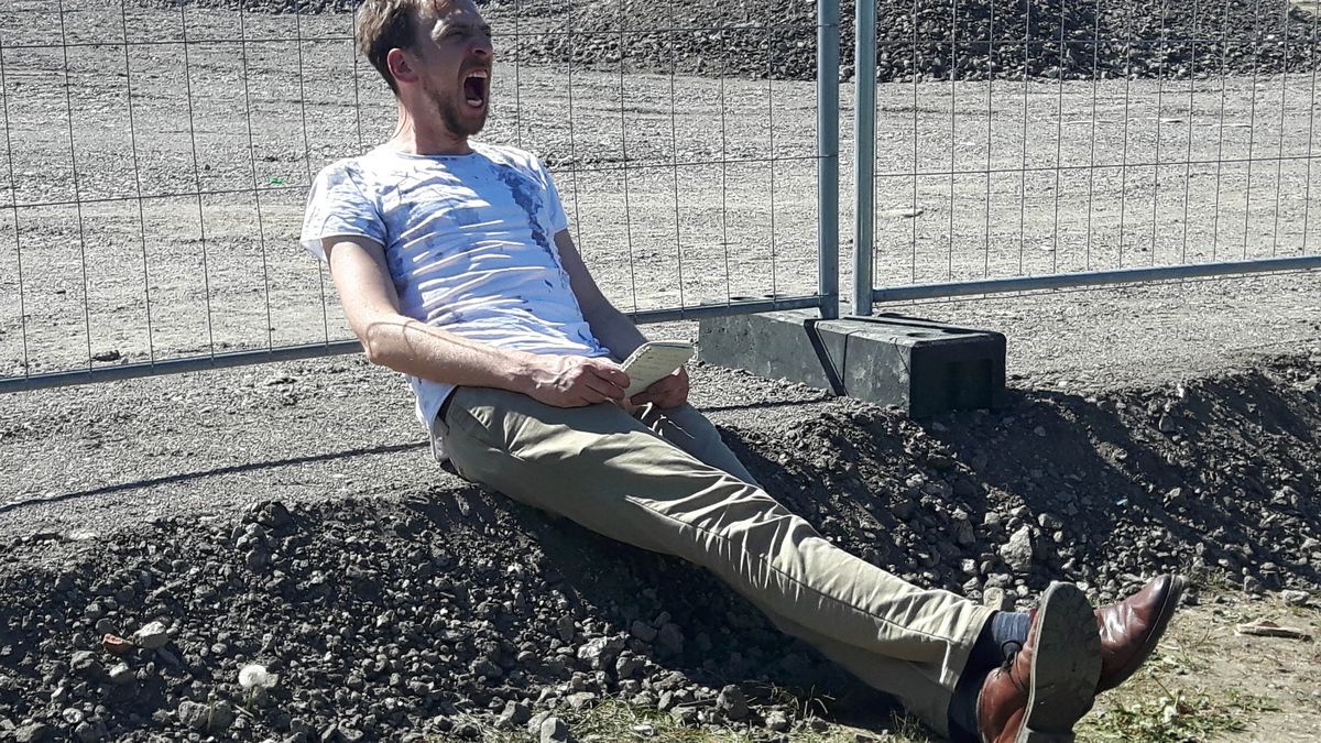 Hermann Heisig lehnt sitzend mit aufgerissenem Mund an einem Bauzaun