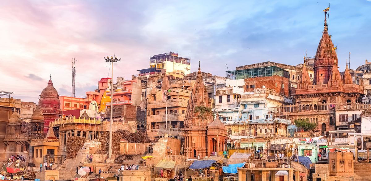 In the center of the picture, you can see the city of Varanasi. In the right foreground, there is a man sitting in a boat. The photo is taken from © Adobe Stock and was shot by Roop Dey. 