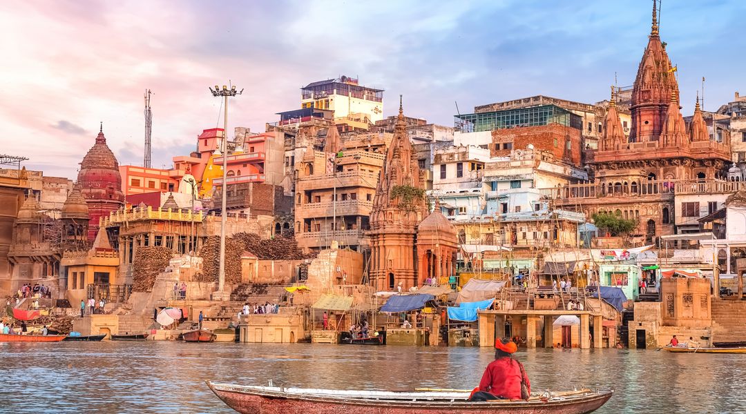 Im Zentrum des Bildes ist die Stadt Varanasi zu sehen. Im rechten Vordergrund sitzt ein Mann in einem Boot. Das Foto kommt von © Adobe Stock und wurde von Roop Dey geschossen. 