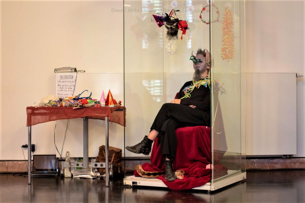 zur Vergrößerungsansicht des Bildes: Eine Studentin sitzt komisch verkleidet in einer Glasvitrine, daneben ein Tisch mit weiteren komischen Sachen