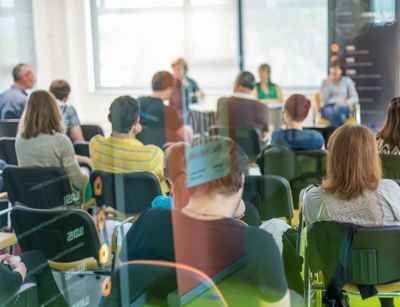 Durch eine Glasscheibe fotografiert sieht man Studierende einer Präsentation folgen