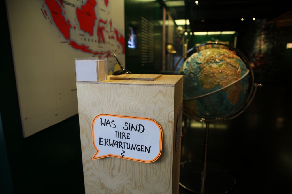 Ein großer Kasten aus Holz steht in einem Museum vor einem Globus. Auf dem Kasten liegen Zettel und Stifte. An dem Kasten hängt eine Sprechblase aus Papier mit der Frage: Was sind Ihre Erwartungen?