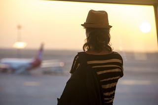 Eine junge Frau mit Hut steht am Fenster und schaut auf das Rollfeld eines Flughafens.