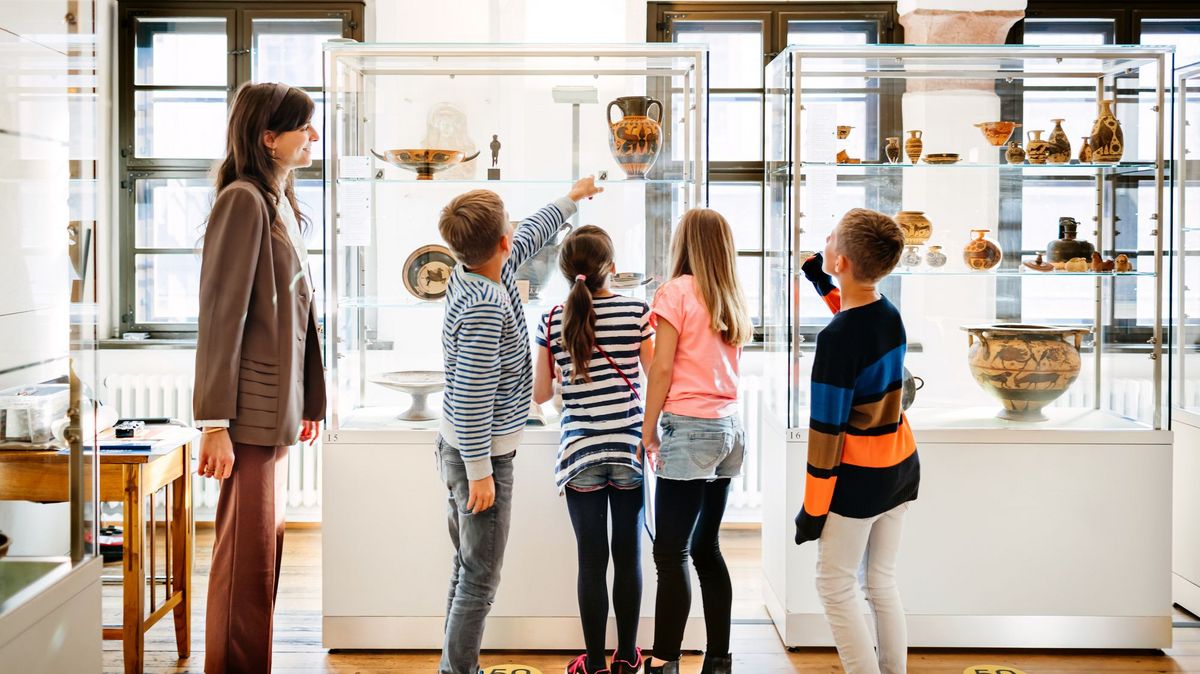zur Vergrößerungsansicht des Bildes: Kinder stehen zusammen mit einer Mitarbeiterin vor einer Vitrine im Antikenmuseum und schauen sich die Ausstellungsstücke an.