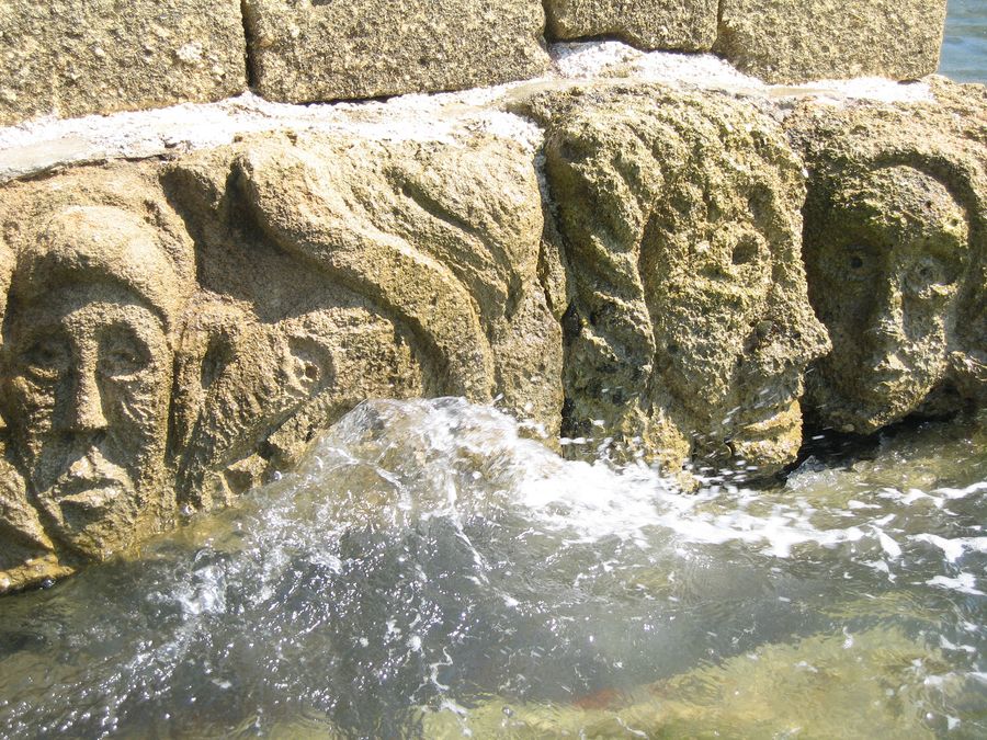 In eine Mauer gehauene Steingesichter, von Meerwasser umspült