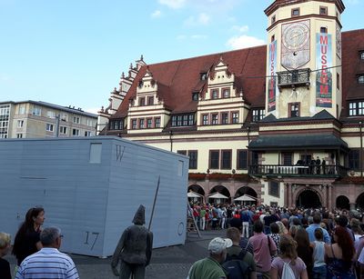 Publikum steht um einen weißen Kubus auf dem Leipziger Marktplatz, den Blick zum alten Rathaus gerichtet