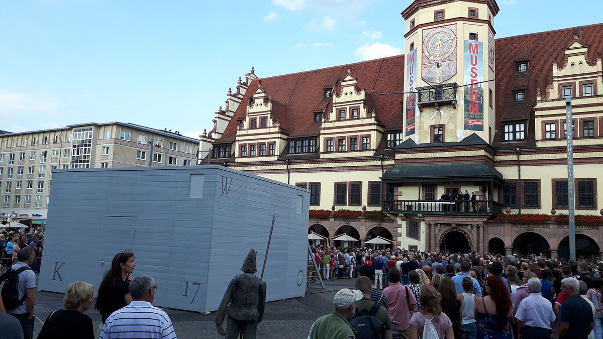 zur Vergrößerungsansicht des Bildes: Publikum steht um einen weißen Kubus auf dem Leipziger Marktplatz, den Blick zum alten Rathaus gerichtet