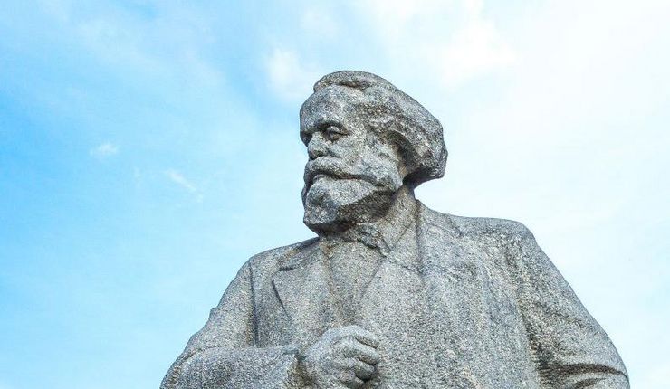 Verwitterte Karl-Marx-Statue vor Bäumen und blauem Himmel. Am Sockel der Statue ist ein russischer Schriftzug eingraviert.