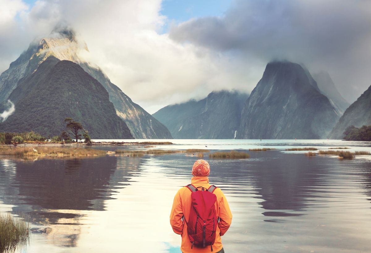 zur Vergrößerungsansicht des Bildes: Mensch steht mit dem Rücken zur Kamera vor einem See und betrachtet diesen und die dahinterliegenden Berge