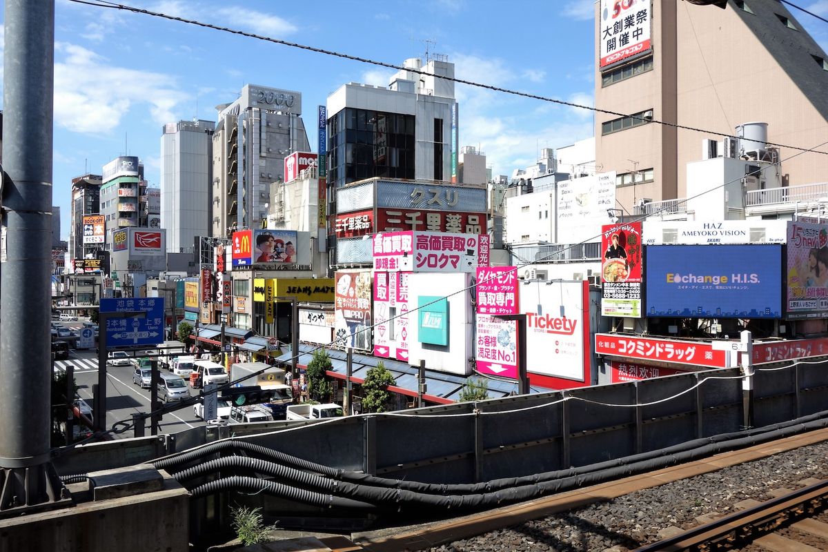 zur Vergrößerungsansicht des Bildes: Das Foto zeigt eine Aufnahme aus der Stadt Tokyo. Im Vordergrund sind Bahnschienen, dahinter zahlreiche Gebäude und eine stark befahrene Straße zu sehen. An allen Gebäuden ist Leuchtwerbung angebracht.