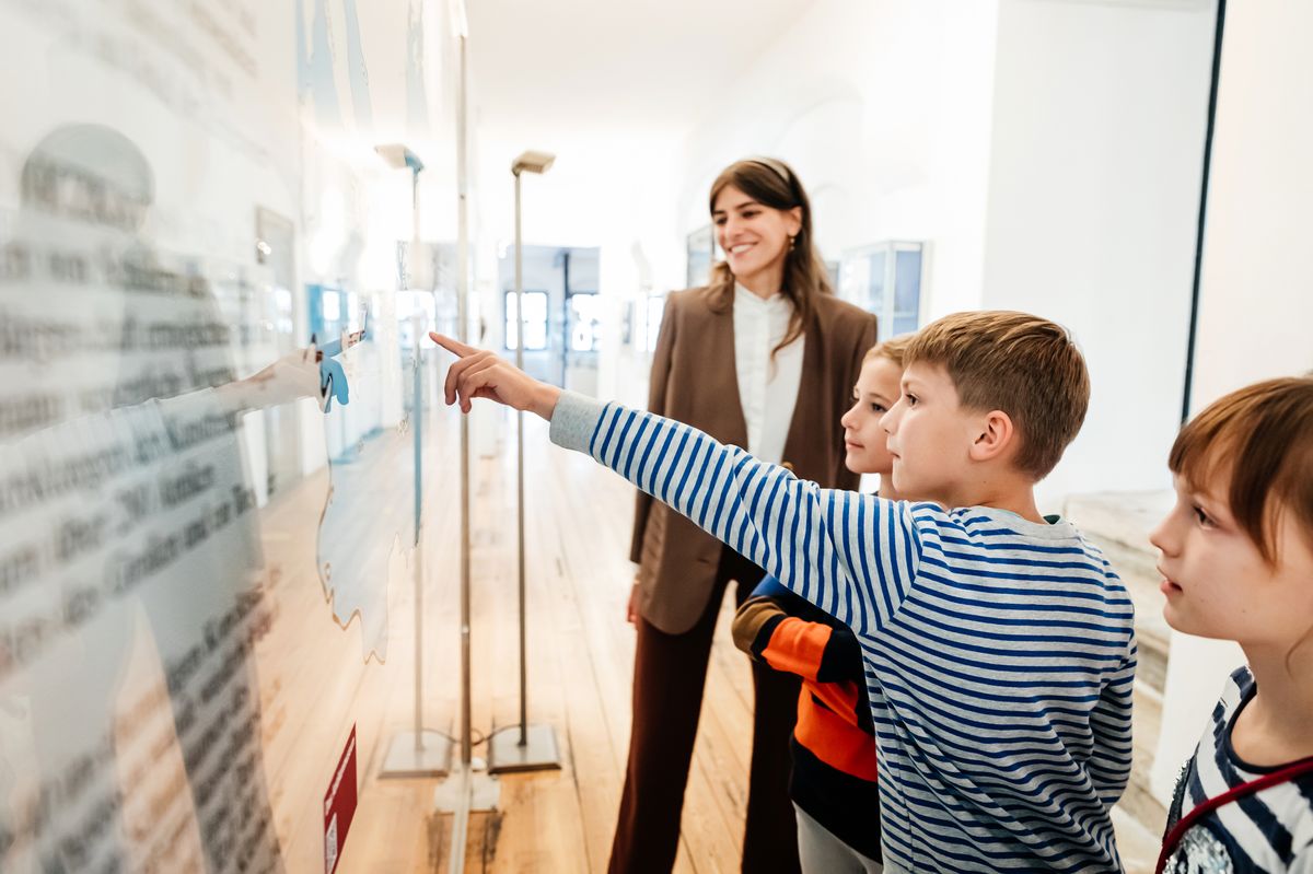 zur Vergrößerungsansicht des Bildes: Kindergruppe und Museumsmitarbeiter stehen im Antikenmuseum vor einer Karte des Mittelmeerraums.