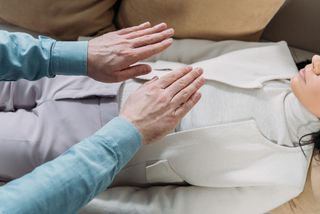 Vogelperspektive: Aufnahme einer liegenden Frau in weißer Kleidung, über ihrer Körpermitte schweben die ausgestreckten Hände eines Therapeuten.